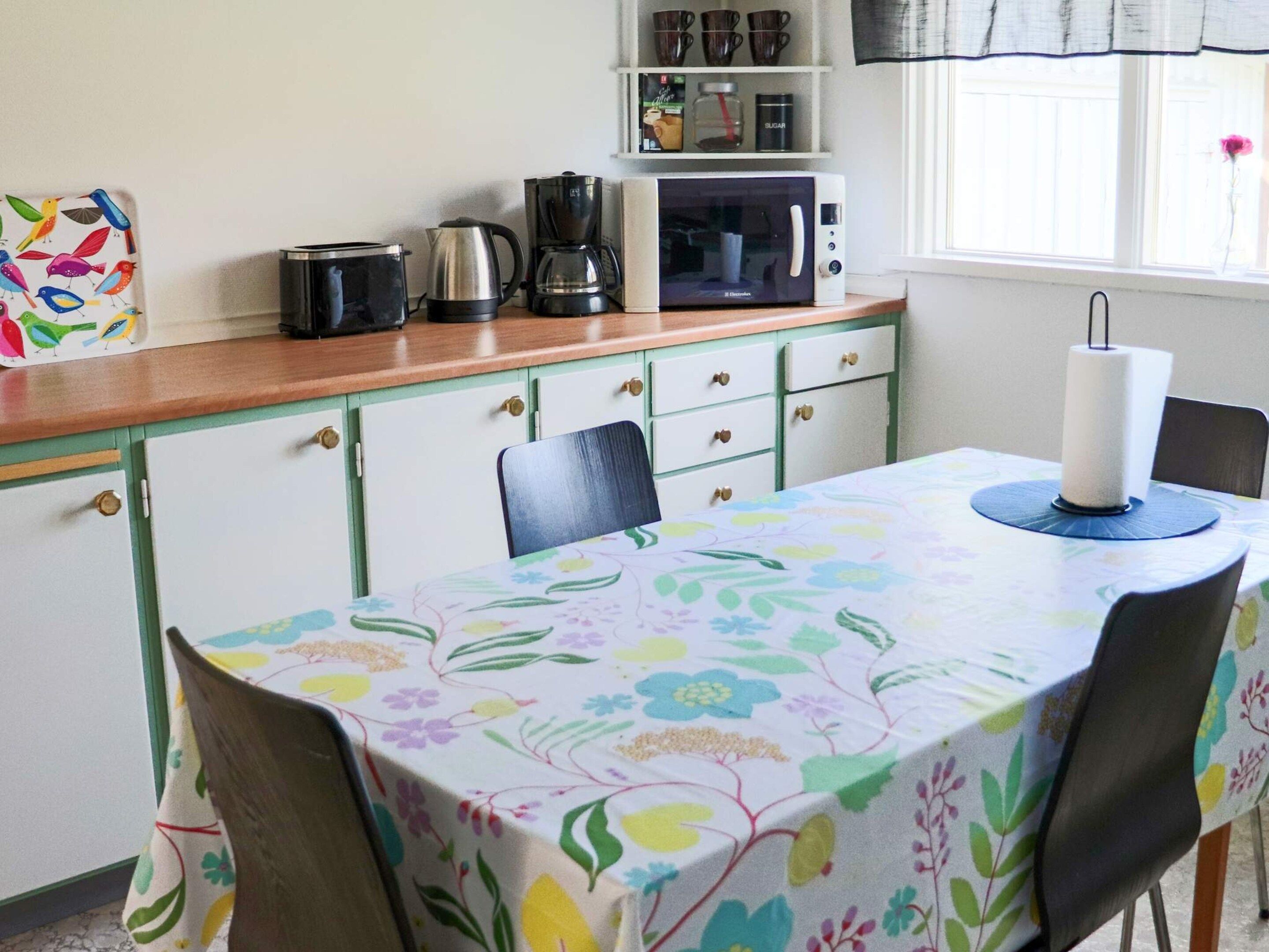 Una cocina con microondas, cafetera y hervidor sobre la encimera. Hay una mesa con mantel floral y una vela.