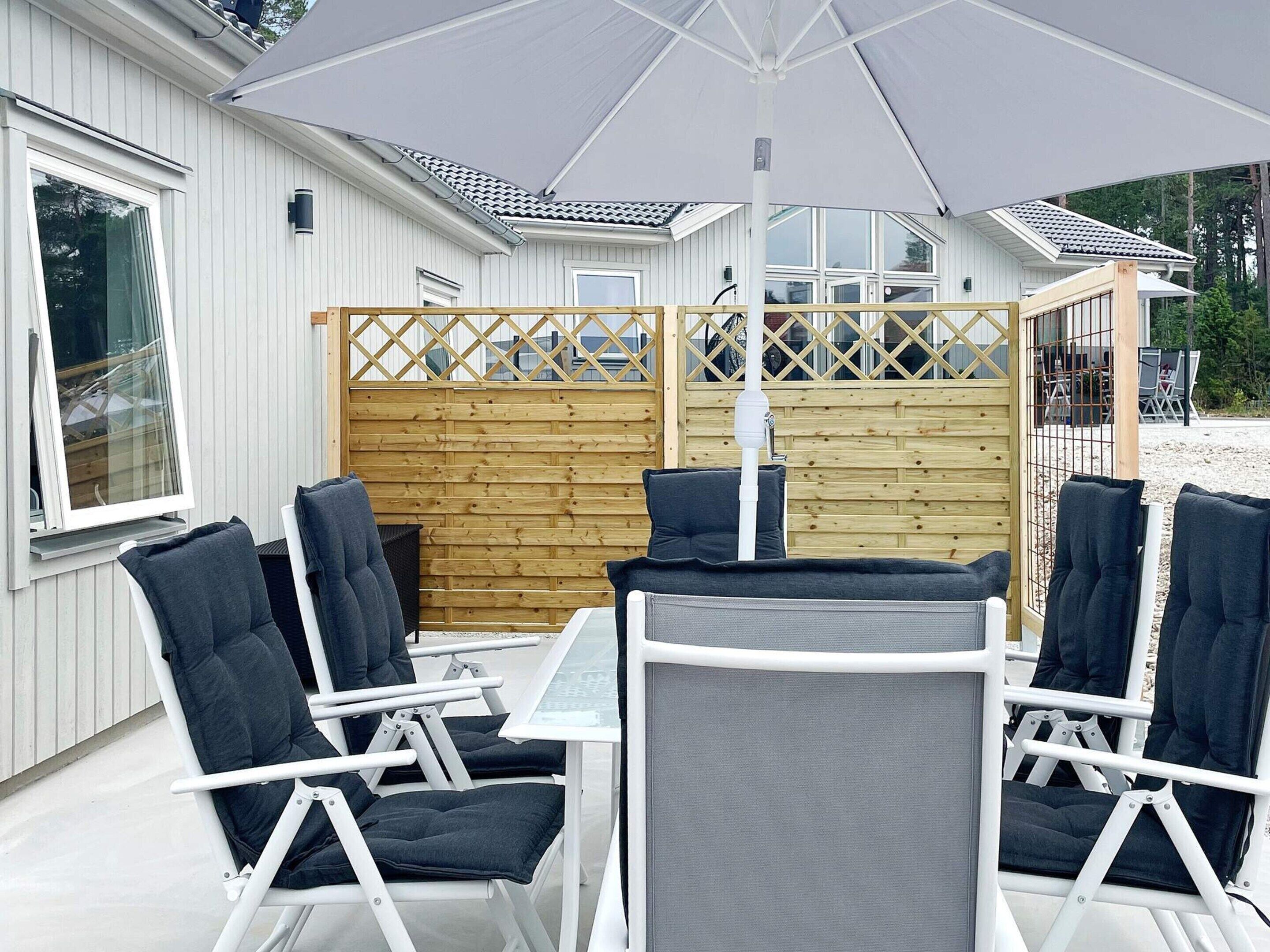 A covered outdoor seating area with white metal chairs and a table, a wooden fence, and a building with large windows in the background.