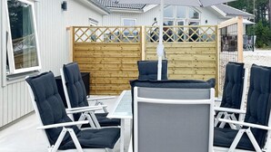Un espacio al aire libre cubierto con sillas de metal blanco y una mesa, una cerca de madera y un edificio con grandes ventanales al fondo.