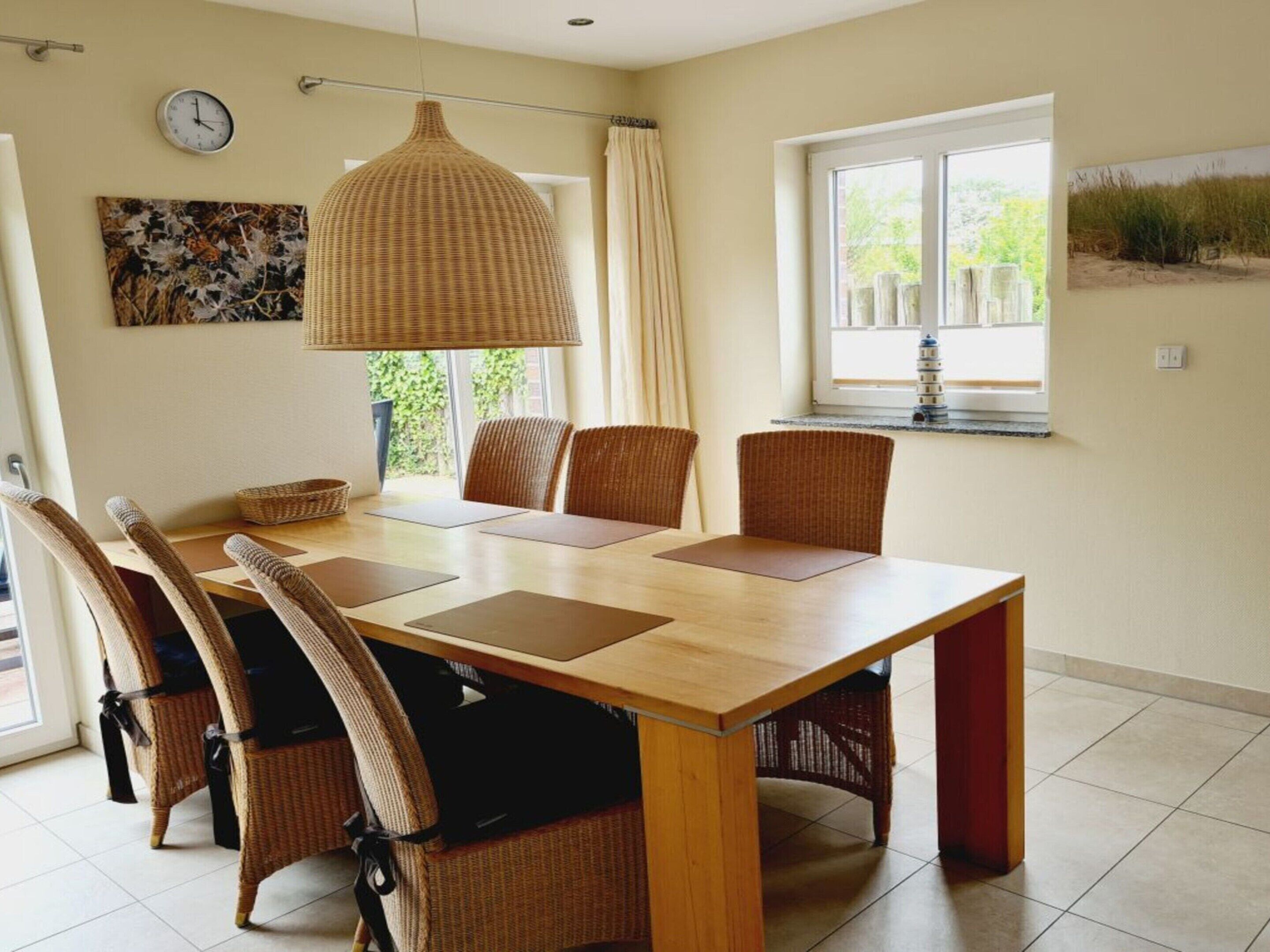Un comedor con mesa de madera, seis sillas de mimbre, lámpara colgante, un reloj y ventana con vista a follaje.