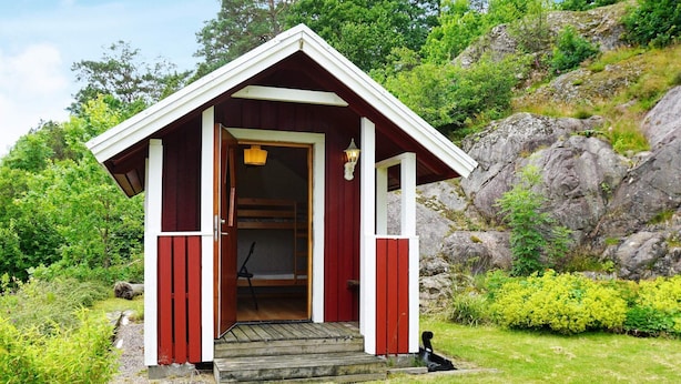 Una cabaña de madera pequeña y roja con una sola puerta y un banco en su interior.
