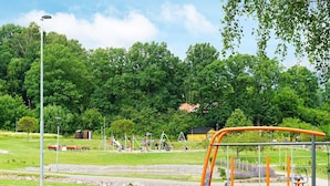 Un parque con un área de juegos, varios equipos, bancos y una mesa de picnic, con árboles y un edificio al fondo.