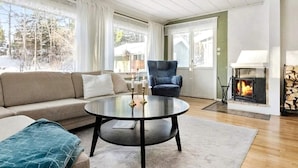 A living room with a sofa, a round coffee table, a blue armchair, a fireplace, and a view of trees outside. - 4 Star Holiday Home in Varmdo (Värmdö)