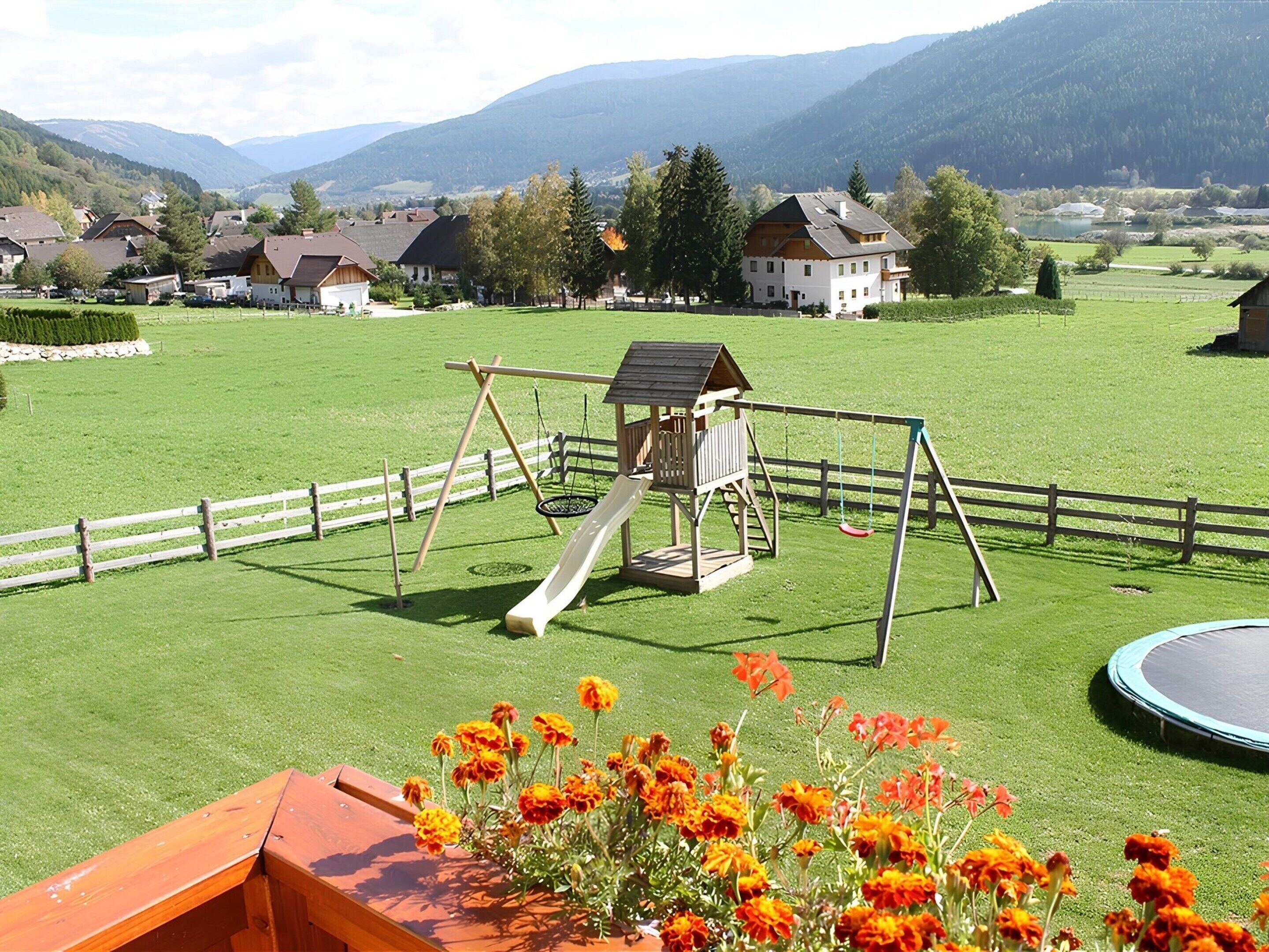A playground with a slide and swings, surrounded by a green field and houses in the background.