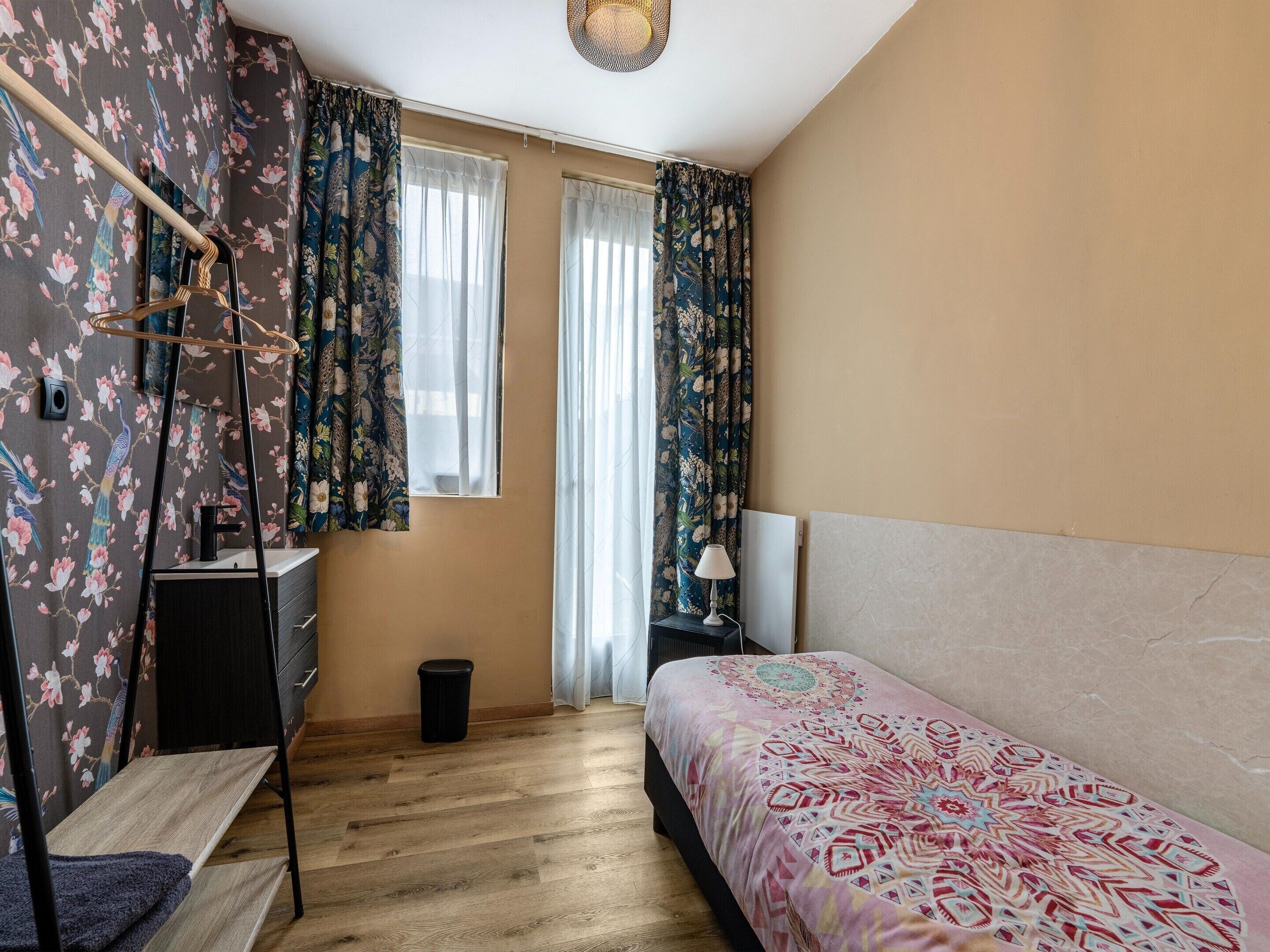 Un dormitorio con cama, una mesita de noche, lámpara, una silla colgante, un cesto de basura y una ventana con cortinas.