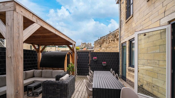 Une terrasse en bois avec un canapé, une table et des chaises, une clôture noire et, au fond, un bâtiment en pierre.