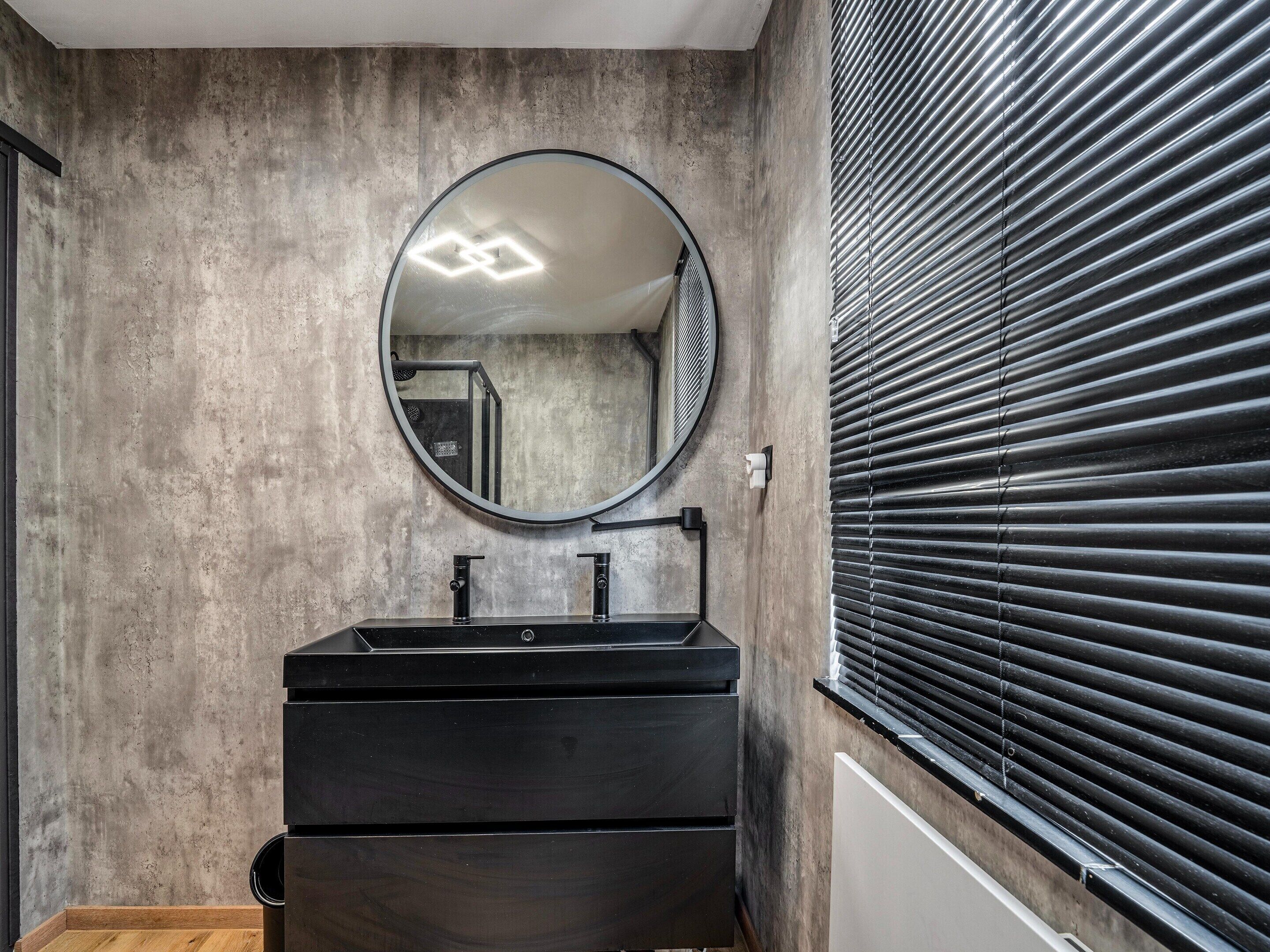 Une salle de bain avec un grand miroir rond, deux lavabos noirs, un plancher en bois et une fenêtre avec des stores.