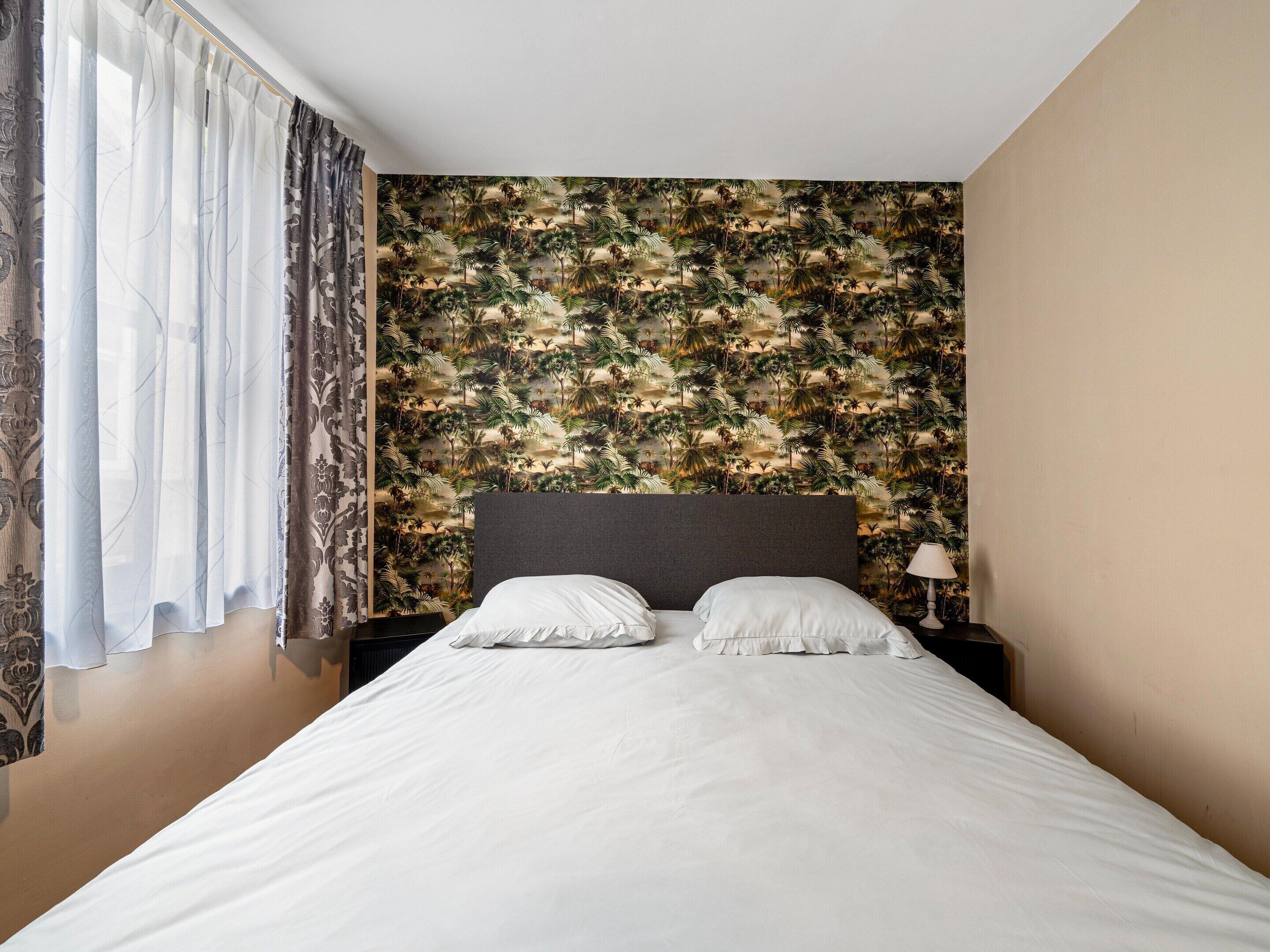 Une chambre d’hôtel avec un lit, deux oreillers, une table de chevet, une lampe et une fenêtre avec des rideaux.