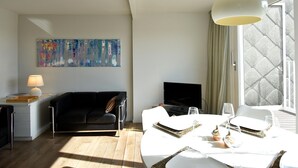 A modern living room with a black sofa, a white table set for dining, a flat-screen TV, and a large abstract painting on the wall. - Bright Flat With Bunk Beds (De Haan)