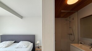 A bedroom with a bed, bedside table, lamp, and a shower area with a glass door and wooden vanity. - Bright Flat With Bunk Beds (De Haan)