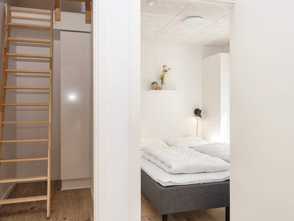 Un dormitorio con una cama, una lámpara de noche, un estante con objetos decorativos y una escalera de madera que da a un armario.