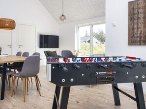 Una habitación moderna con una mesa de futbolín, un comedor con sillas y una mesa, un televisor montado en la pared y una ventana con vistas al exterior.