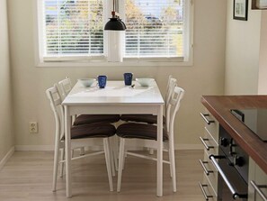 Un coin repas avec une table et des chaises blanches, un luminaire suspendu et une fenêtre avec des stores.