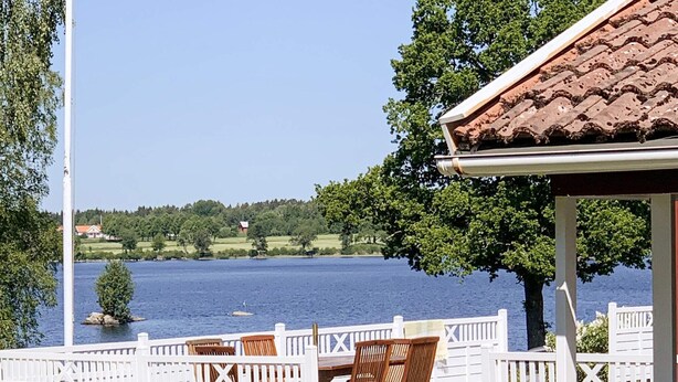 A covered outdoor seating area with wooden chairs and a grill, overlooking a lake with a white fence and trees. - 4 Person Holiday Home in Herrljunga-by Traum (Herrljunga)