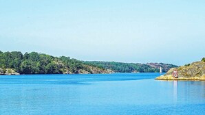 Un lago tranquilo con cielo despejado, rodeado de un bosque verde frondoso y terreno rocoso.
