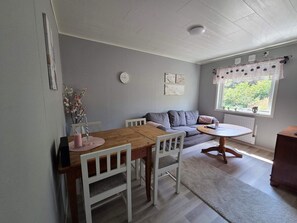 Una sala de estar acogedora con una mesa de comedor de madera, sillas blancas, un sofá gris y una pequeña mesa redonda con mantel blanco.