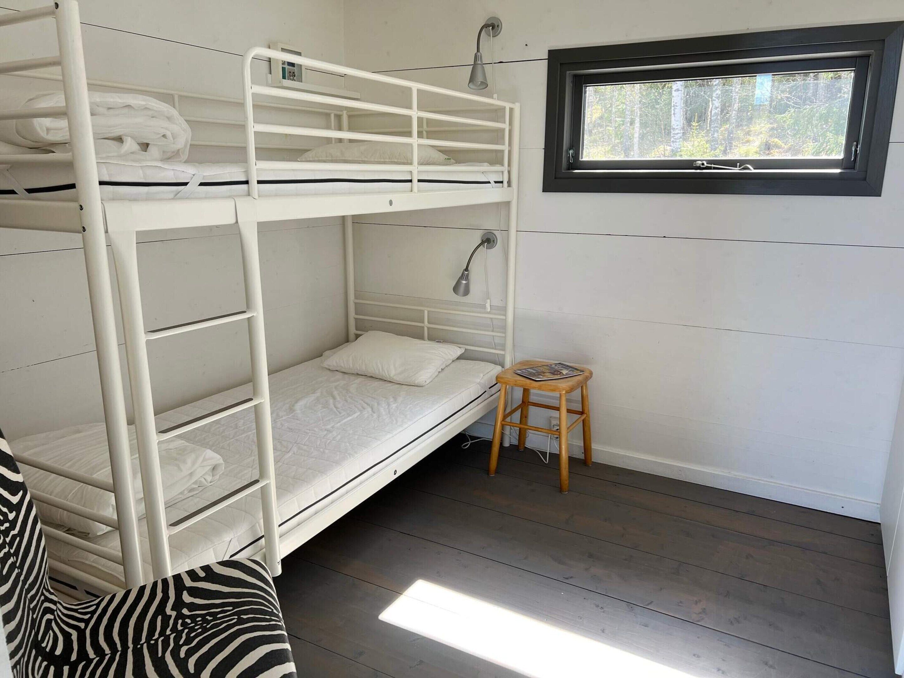 A bunk bed room with a window, a bedside lamp, and a small wooden stool.