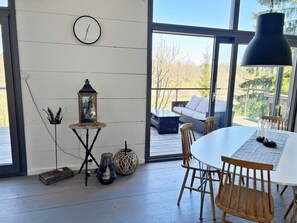 En modern inredning med ett runt matbord, trästolar, ett soffbord i glas, en väggklocka och utsikt över utemiljön genom stora fönster.