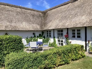 Un cottage dal tetto di paglia con un piccolo patio, pareti bianche e un giardino con cespugli e un tavolo con sedie.