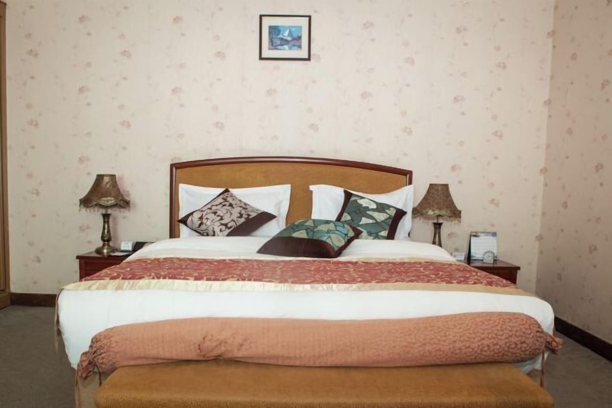 Een net opgemaakt bed met een houten hoofdeinde, twee nachtkastjes met lampen, en een ingelijste foto aan de muur.