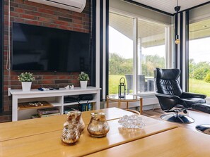 A modern living room with a flat-screen TV mounted on a brick wall, a wooden table with decorative items, a black leather chair, and a view of a garden through large windows. - 6 Person Holiday Home in Brenderup Fyn (Middelfart)