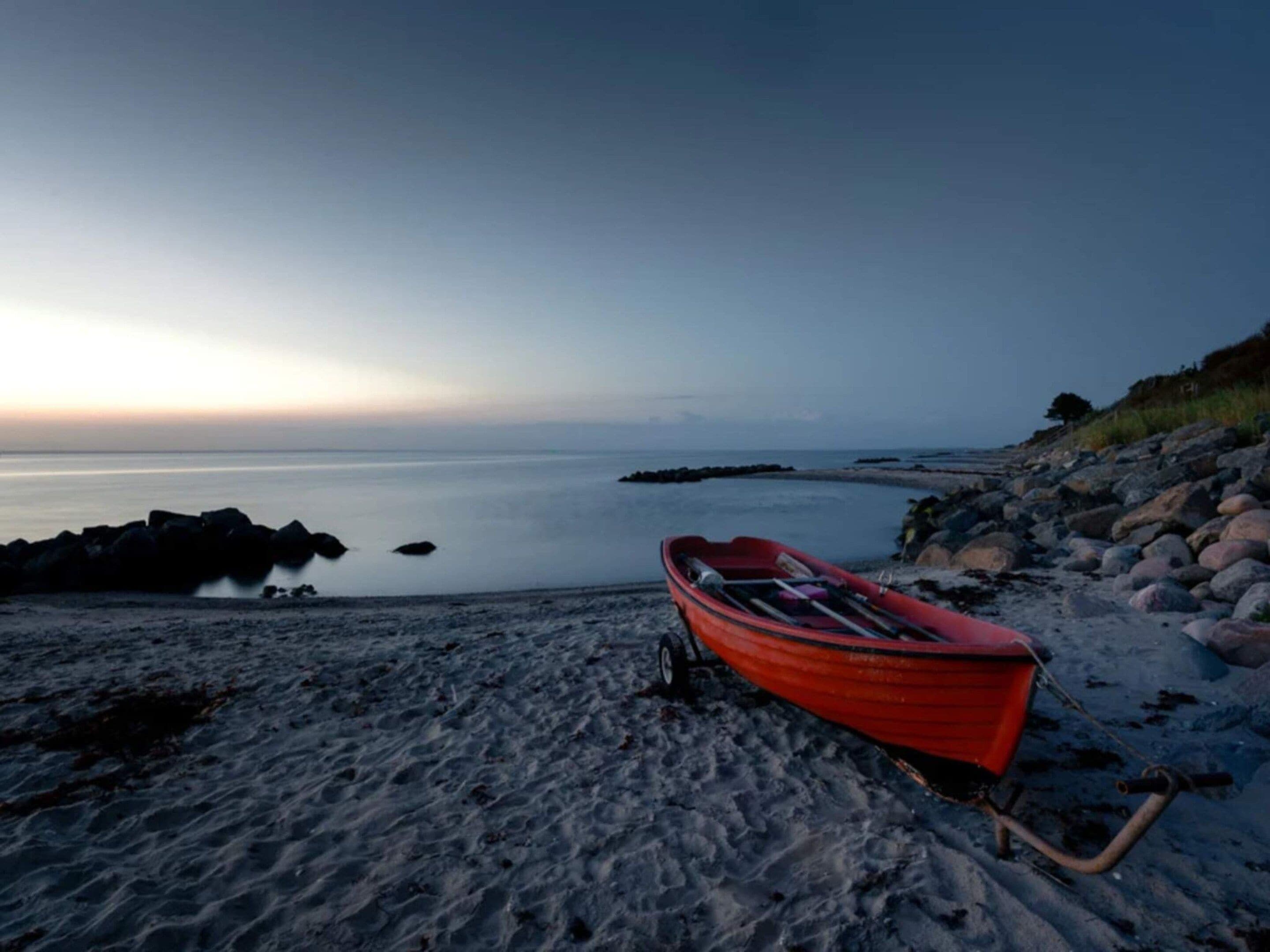 A red boat on a sandy beach with a calm sea and rocky shoreline.