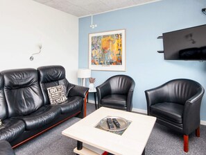 Ein Zimmer mit einer schwarzen Ledersofa, zwei schwarzen Ledersesseln, einem kleinen weißen Tisch mit einer Fernbedienung, einem gerahmten abstrakten Gemälde, einem wandmontierten Fernseher und einer Lampe.