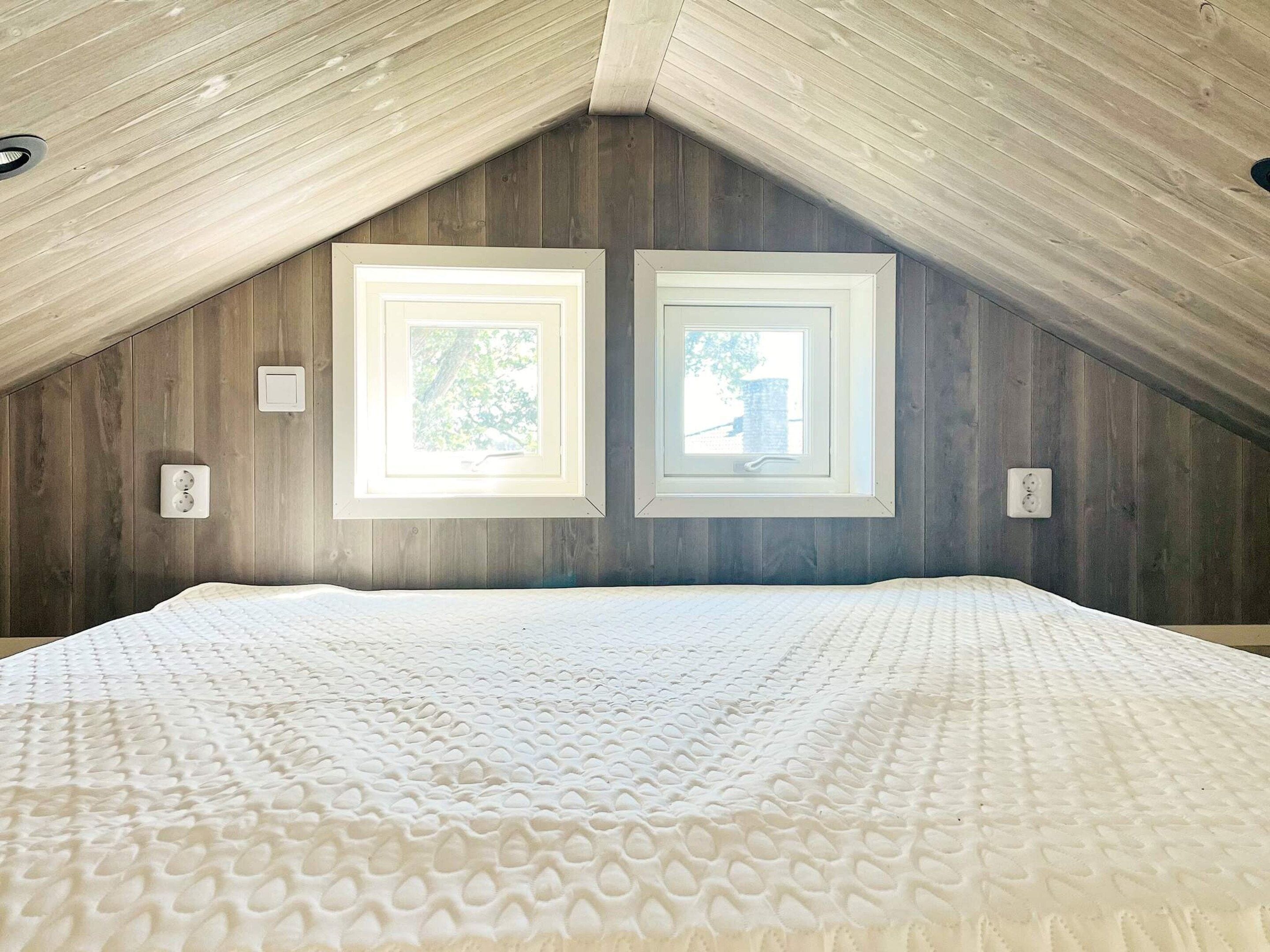 A bedroom with a sloped ceiling, two windows, a bed with a textured mattress, and wooden walls.