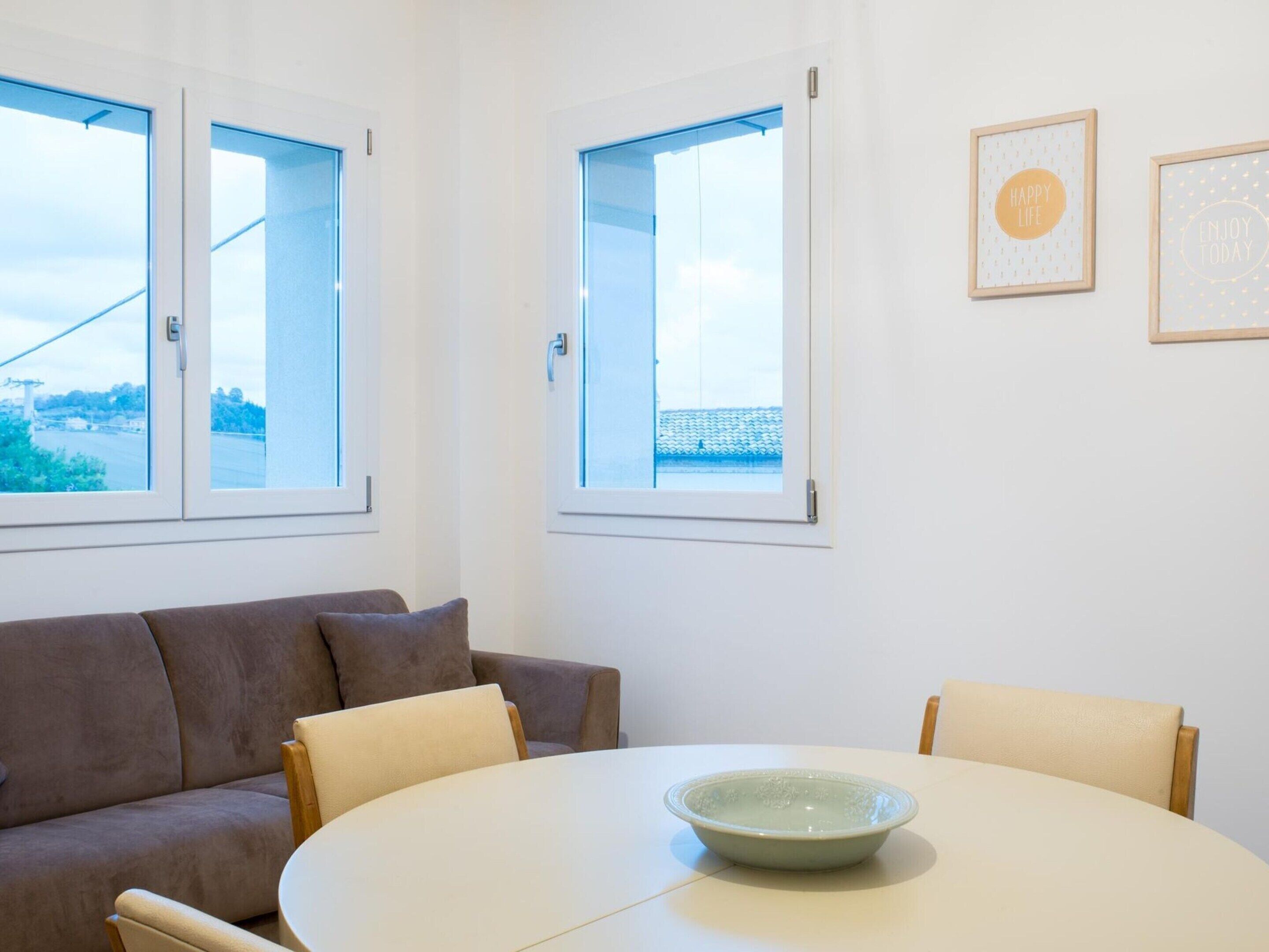 Habitació amb una taula rodona, dues cadires, un sofà i dues finestres que donen vistes a un edifici blau i vegetació.