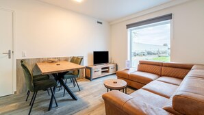 Un salon moderne avec une table à manger en bois, des chaises vertes, un canapé en cuir, un téléviseur et une grande fenêtre donnant sur l’extérieur.