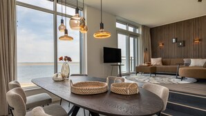 A modern living room with a large dining table, comfortable seating, a flat-screen TV, and a view of the sea.