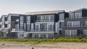 A modern multi-story residential building with balconies, a sandy beach in the foreground, and a clear sky.