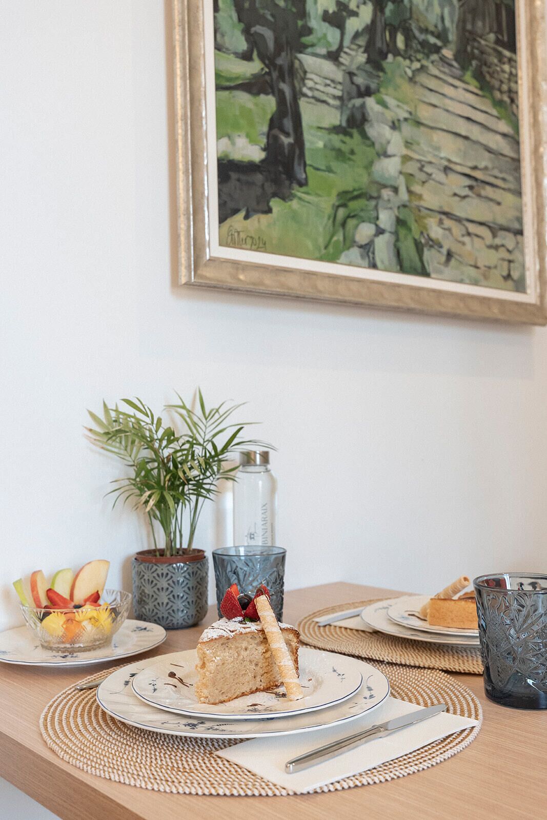 A table with a slice of cake on a plate, a glass of water, a potted plant, and a framed painting on the wall.