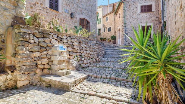 Calle empedrada y estrecha bordeada por edificios tradicionales de piedra, con pequeñas ventanas y un muro de piedra con escaleras que bajan.