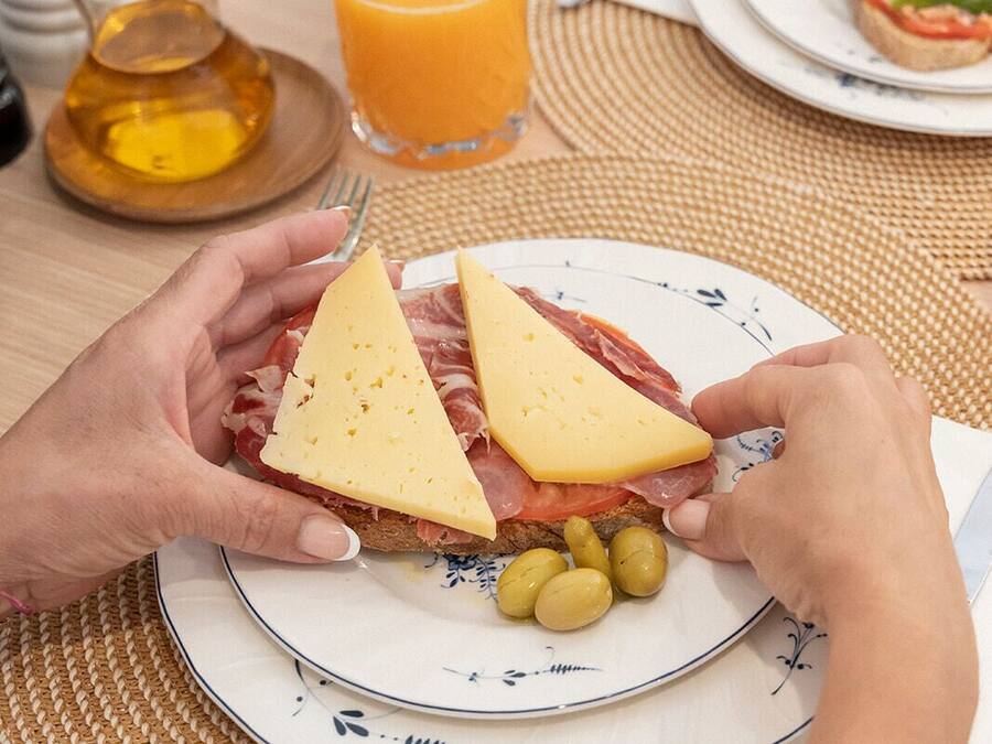 Una persona sosteniendo un sándwich de queso y jamón, un vaso de jugo de naranja y aceitunas en un plato.