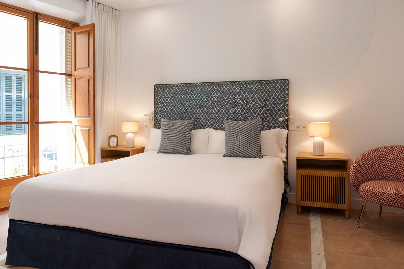 A hotel room with a large bed, two bedside tables with lamps, a patterned headboard, a red patterned chair, and a window with curtains.