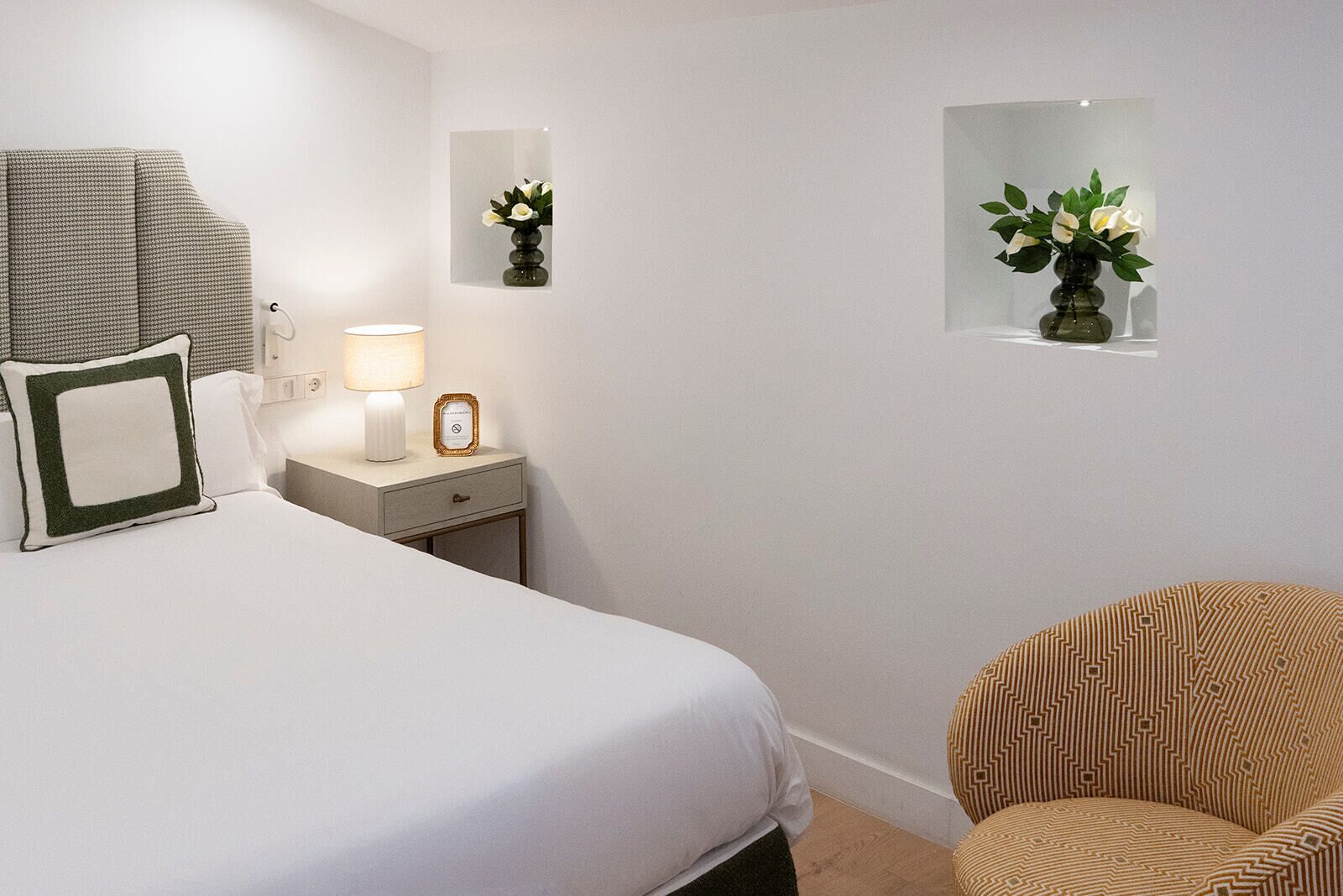 Habitación de hotel con una cama, una mesita de noche con lámpara, una silla y dos espejos adosados con jarrones de flores.