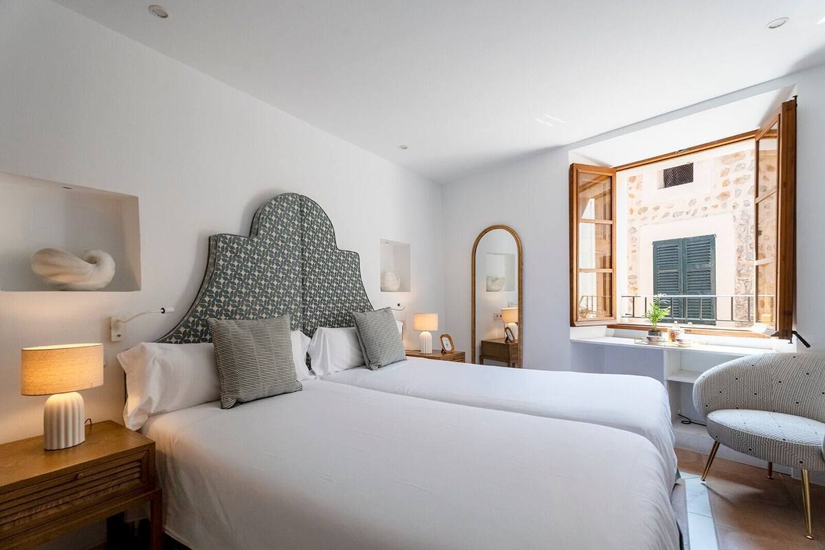 A hotel room with two beds, a sitting area with a chair, a window with a view of a building, and a small table with a lamp.