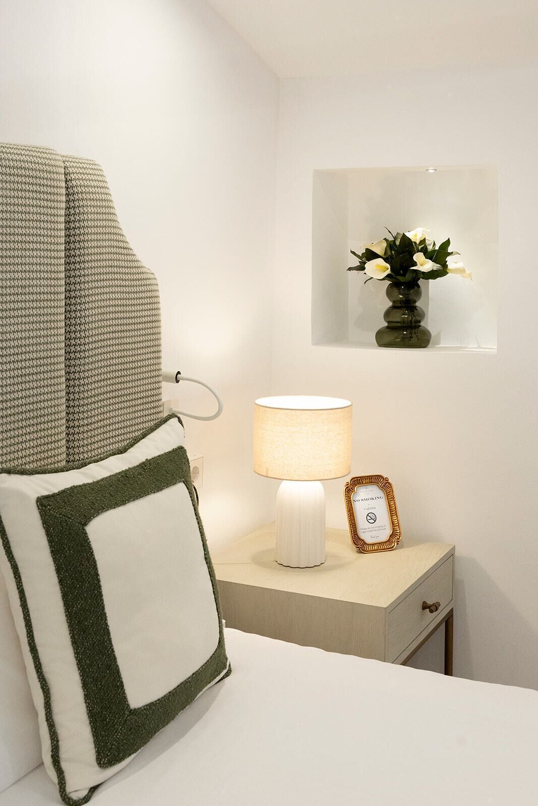 A neatly arranged bedroom with a bed, a bedside lamp, a small bedside table with a framed sign, and a wall-mounted mirror with a vase of flowers.