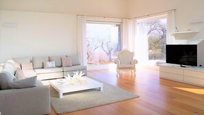 A modern living room with a sofa, coffee table, and armchair, a flat-screen TV, and a large window with a view of trees. - The Great Beauty, beautiful property with amazing view on the lake of Como (Lombardia)
