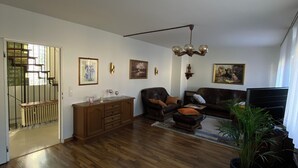 A spacious living room with wooden flooring, a brown leather sofa, a coffee table, and a potted plant. There is a staircase leading to an upper floor, and the room has a ceiling fan. - Ferienhaus Eschborn, Frankfurt, Messe (Eschborn)