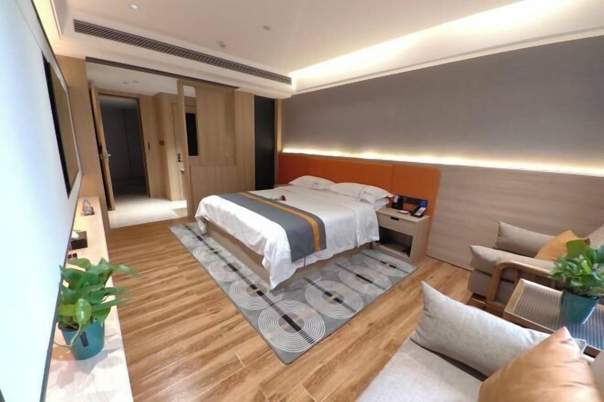Una habitación de hotel moderna con una cama grande, una mesita de noche con lámpara, un rincón de estar con un sillón y un sofá, y una planta en maceta.