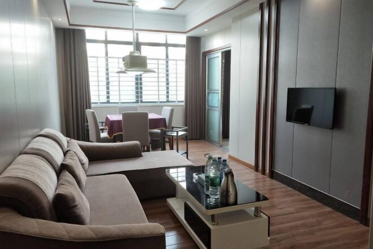 Una habitación de hotel moderna con sofá, una mesa de centro con botellas de agua, televisor montado en la pared y un comedor con mesa y sillas.