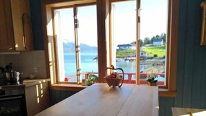 A kitchen with a large wooden table, a window with a view of a lake and houses, and a basket with fruits on the windowsill. - Charming sea house with private sauna & jetty with direct access to the fjord (Vestland)