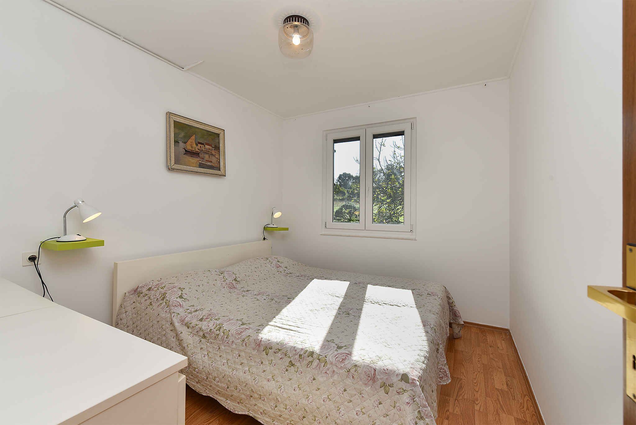 A bedroom with a bed, a desk, a window, and a painting on the wall.