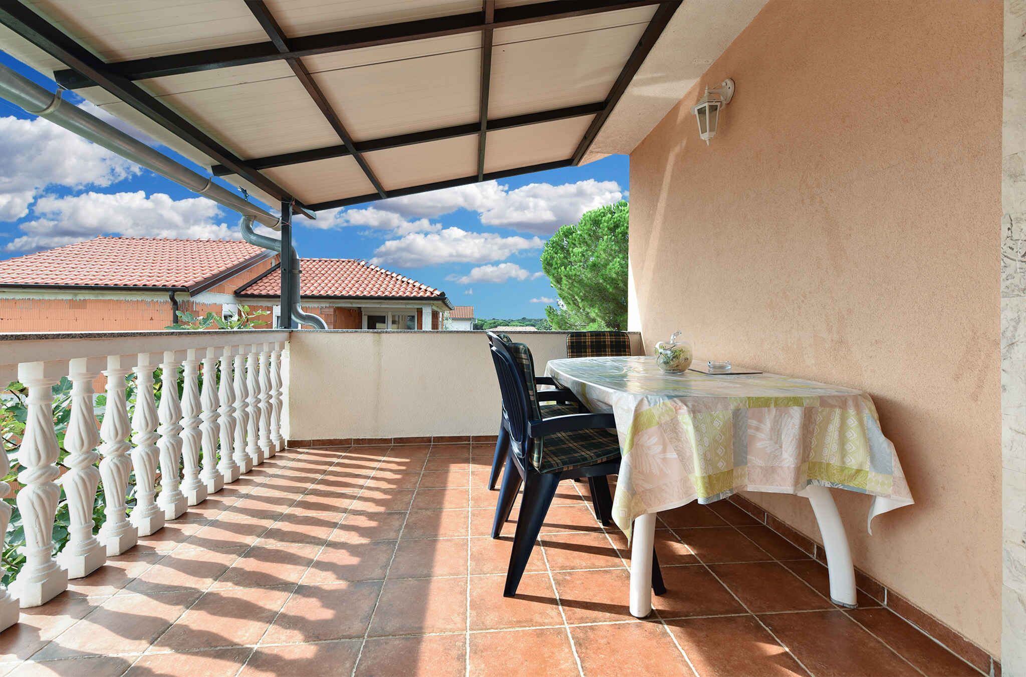 Um pátio coberto ao ar livre com uma mesa e cadeiras, um corrimão branco e vista para edifícios ao fundo.
