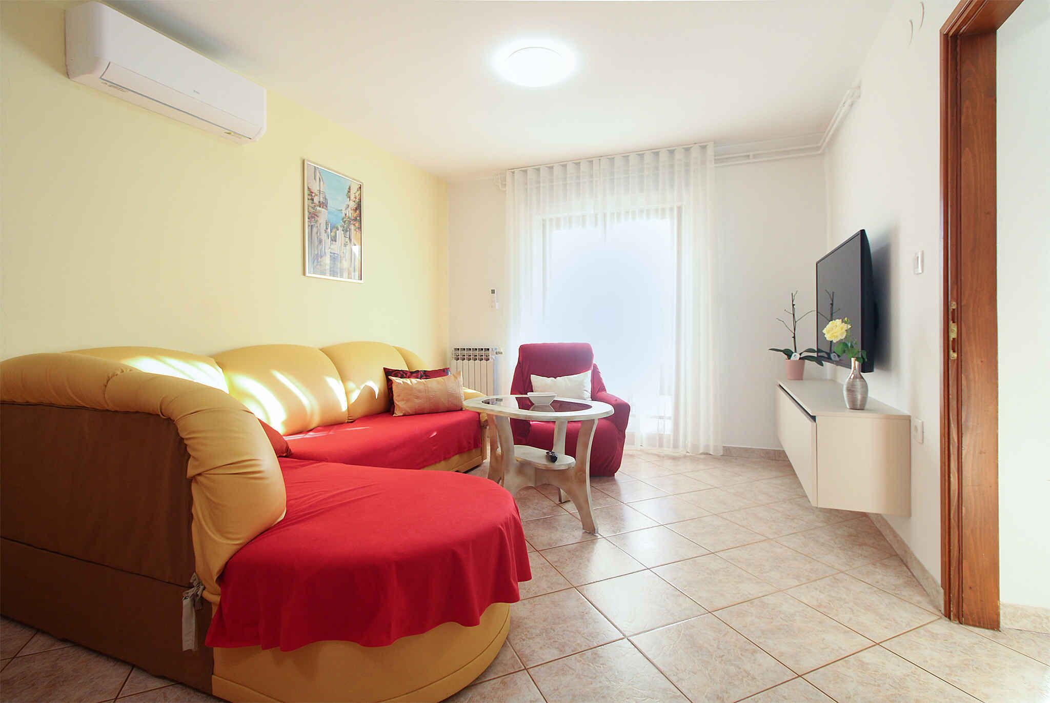 Ein Wohnzimmer mit Sofa, kleinem Tisch, Sessel, Fernseher und einem Fenster mit Vorhängen.