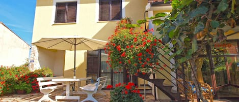 A two-story house with a patio area featuring a table and chairs, a potted plant with red flowers, and a staircase leading to the upper floor.