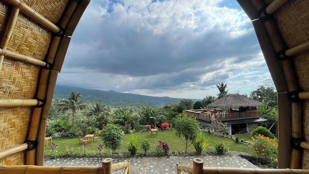 A view from a bamboo-framed window overlooking a lush garden with a thatched-roof building. - Villa Bambu Rinjani & Restaurant By Zuzu (Bayan)