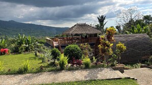 A thatched-roof structure with a wooden deck and a garden with various plants and flowers. - Villa Bambu Rinjani & Restaurant By Zuzu (Bayan)